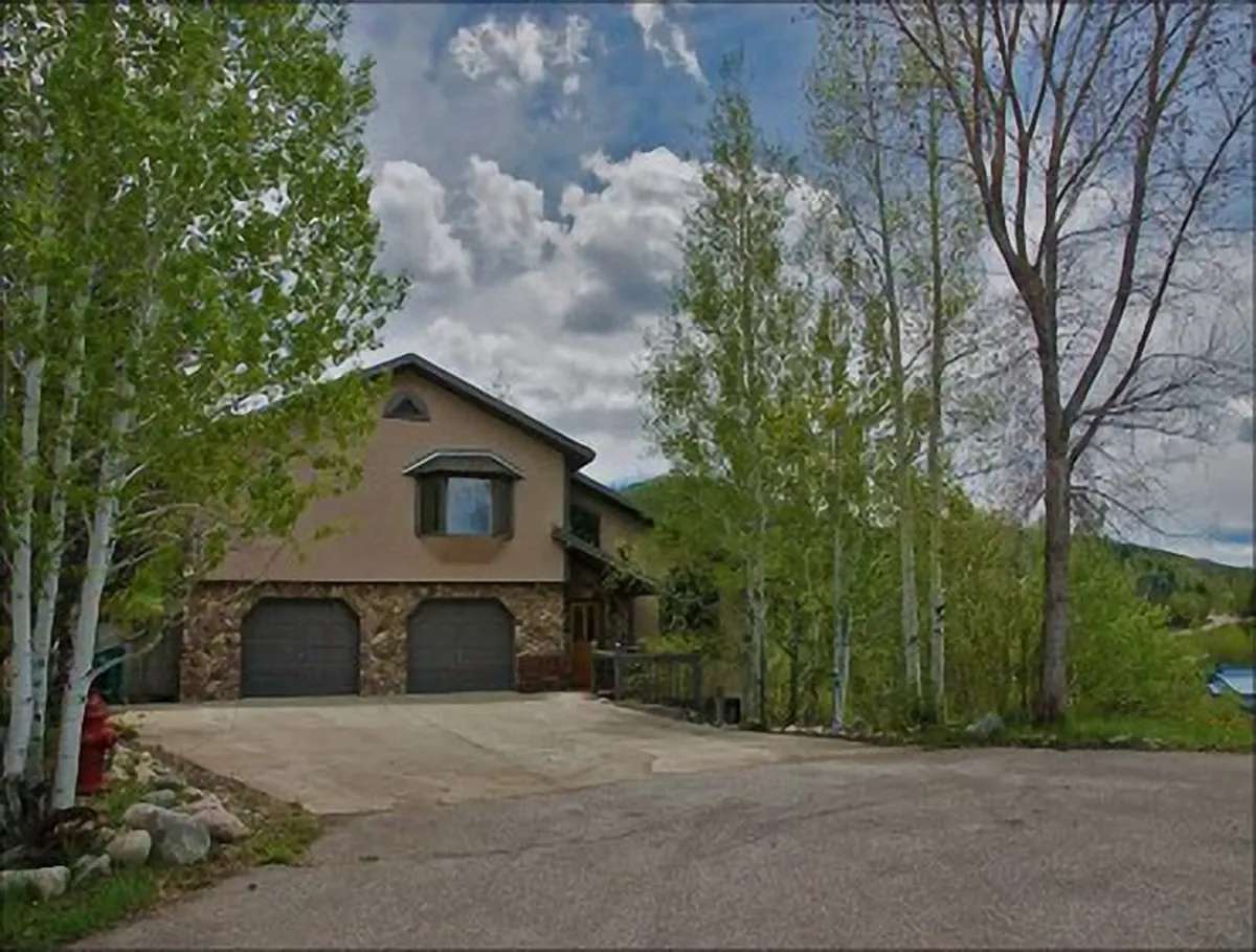 This exterior image shows the home's welcoming facade surrounded by mature trees, with garage doors and open outdoor spaces visible. The landscape sets the scene for gatherings and nature appreciation, and hints at the easy access to and from the property after a day on the slopes.