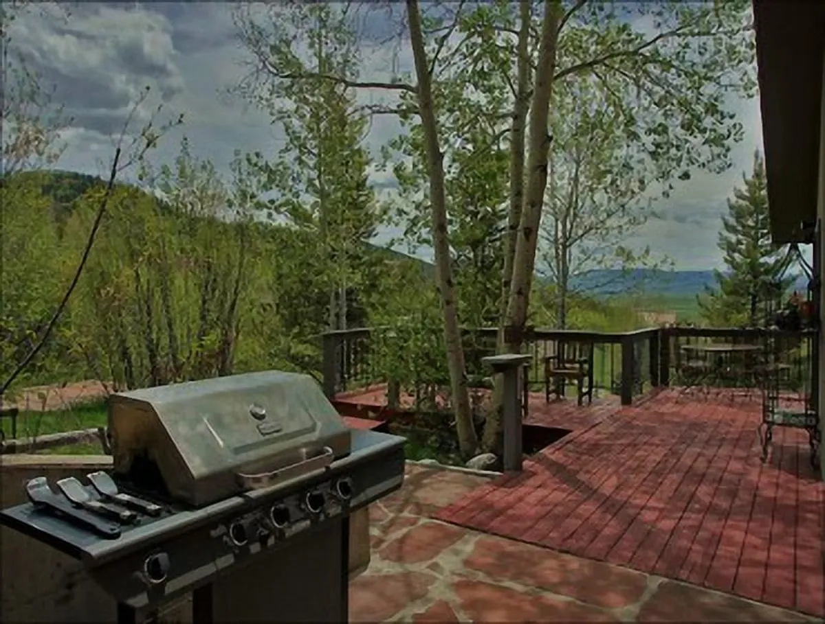This image features a wooden deck outfitted for outdoor living, including a grill and seating, surrounded by lush trees and offering a clear view of the mountains in the distance. The setting is ideal for relaxing, dining, or simply soaking in natural beauty, especially during sunset or sunrise.