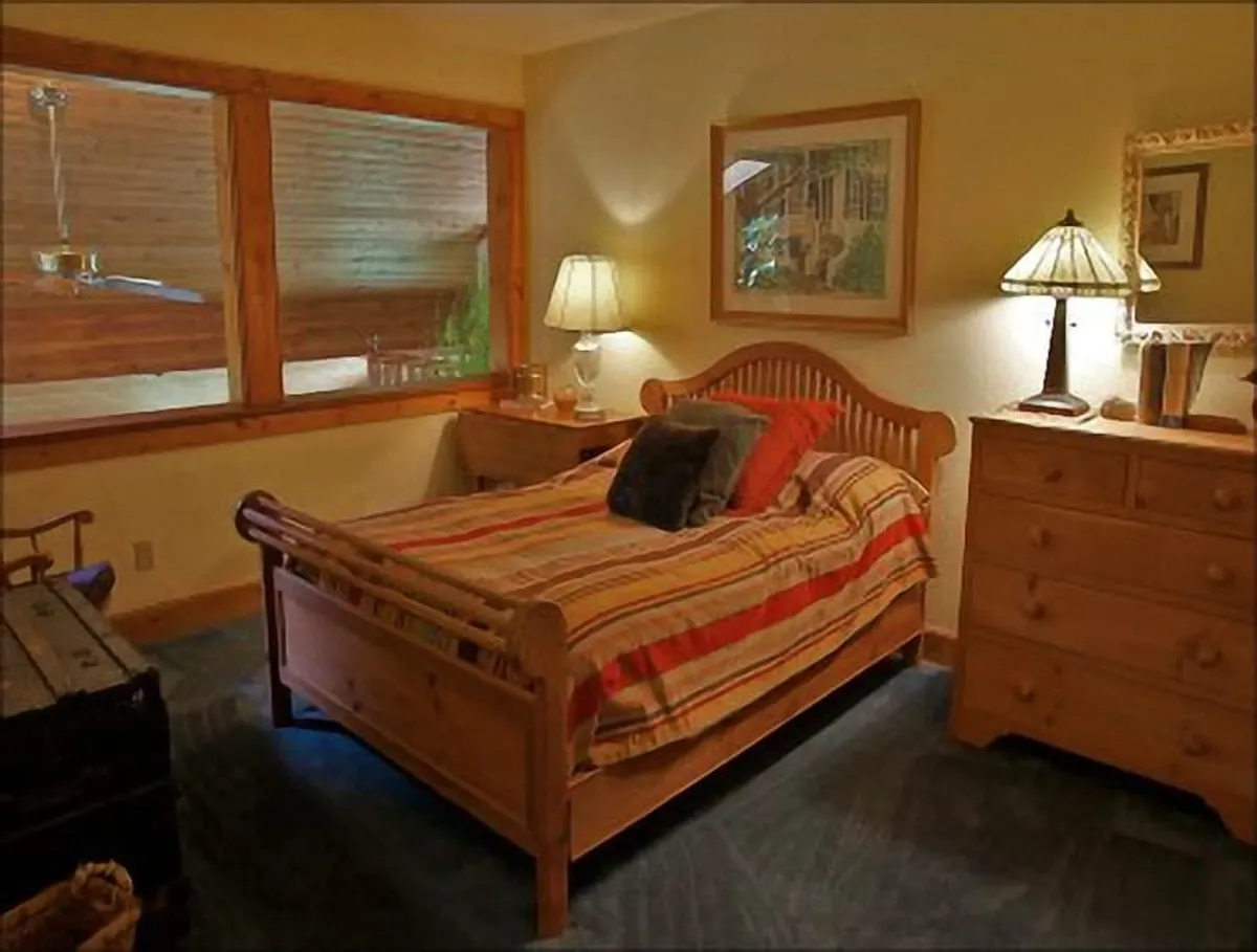 This inviting bedroom features a bed with striped bedding, two bedside lamps, a wooden dresser, and framed artwork. Sunlight filters through the window, highlighting a calm and cozy spot ideal for quiet moments and restful nights.