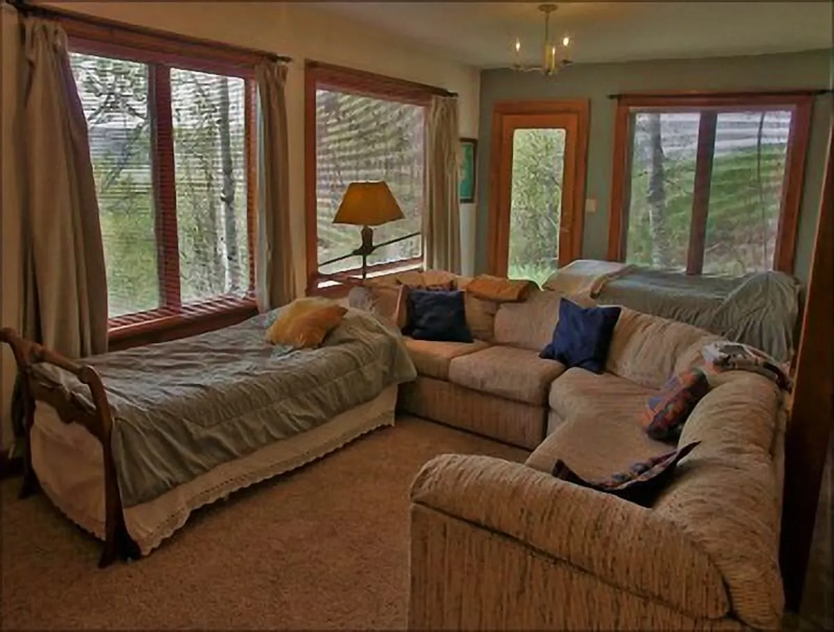 A spacious living area with a large sectional sofa, a bed, a lamp, and expansive windows showcasing views of the natural landscape. The inviting seating arrangement and abundant natural light convey a comfortable setting for unwinding and enjoying time together after a day outdoors.