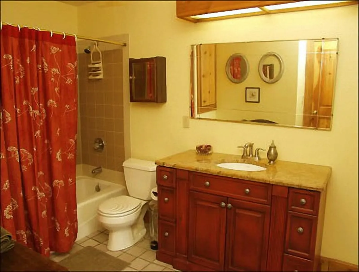 A modern bathroom with a warm, inviting vibe, featuring a wooden vanity with a granite countertop, a mirror with decorative frames, and a bold red patterned shower curtain. The space appears functional and stylish, ideal for unwinding or morning routines.