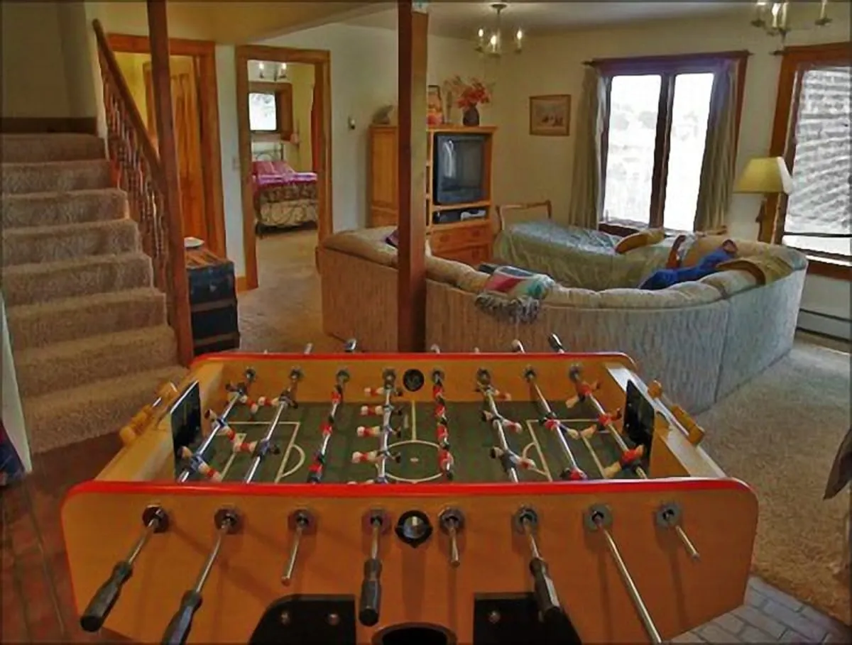 A fun and relaxed living space with a foosball table, sofa, television, staircase, and large windows letting in plenty of light. The setup is ideal for movie nights, friendly games, or letting kids and teens have their own hangout zone.