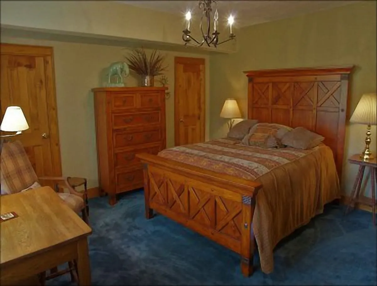 A warmly lit bedroom showcasing a wooden bed frame, chest of drawers, a table lamp, and a decorative plant. The space feels inviting and tranquil, emphasizing relaxation and comfort for guests.