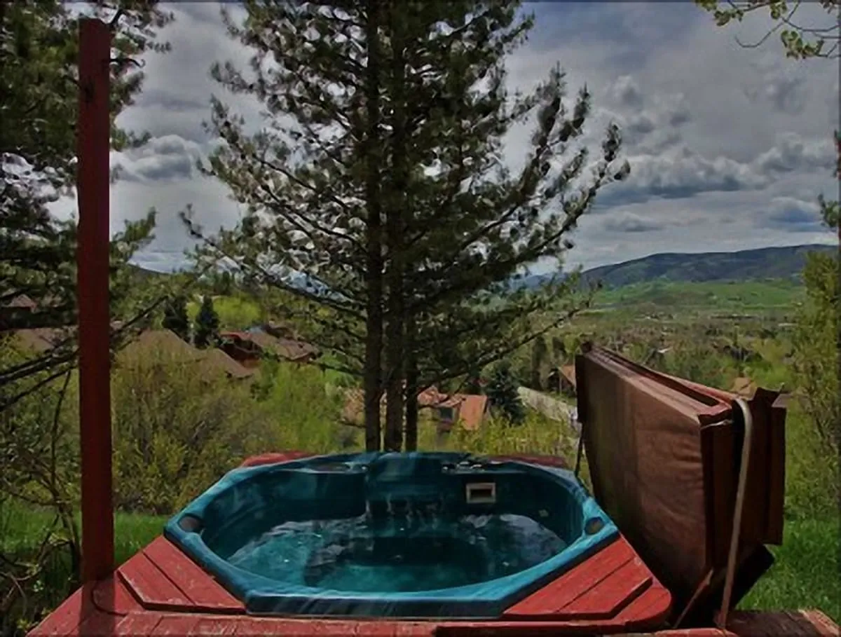 The image features the exterior of a spacious single-family home nestled among towering trees, with a private hot tub overlooking sweeping mountain views. The serene setting invites relaxation, with outdoor seating perfect for soaking in the surroundings, sharing laughs, or gazing at starlit skies.