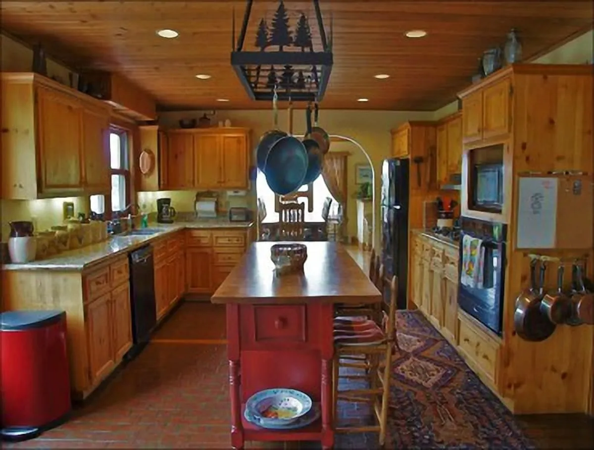 This image showcases a warm, welcoming kitchen with a central island, hanging pots, and wooden cabinetry. A cozy rug grounds the space, and the overall atmosphere is inviting. The scene is set for group cooking, conversation, and shared meals, emphasizing the kitchen as the home's heart.