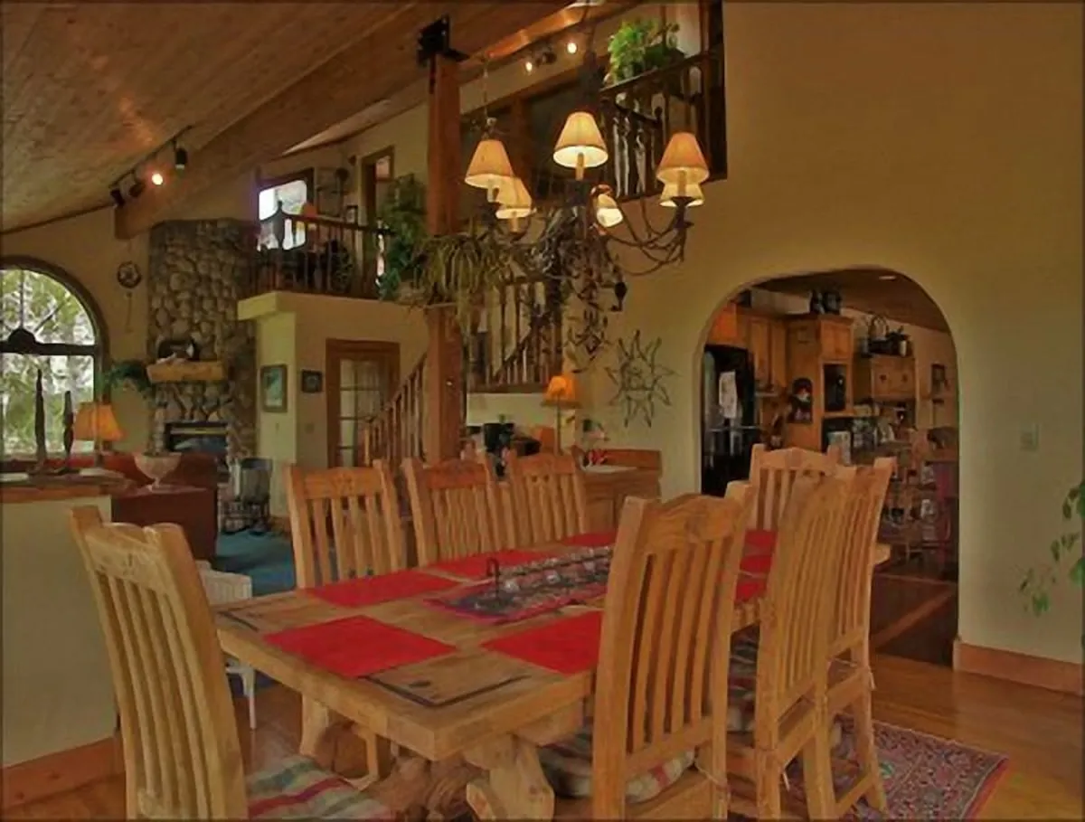 The image displays a large wooden dining table beneath a chandelier, with a stone fireplace and an arched window adding warmth to the scene. The space is set for communal dinners, celebrations, and laughter-filled evenings, perfectly suiting the section’s focus on group gatherings.