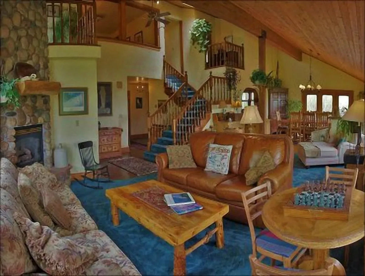 A spacious living area featuring a large leather sofa arranged for group seating, a striking stone fireplace, a wooden dining table set to the side, and a visually interesting spiral staircase. The scene feels lively and family-friendly, perfect for both gatherings and relaxation.