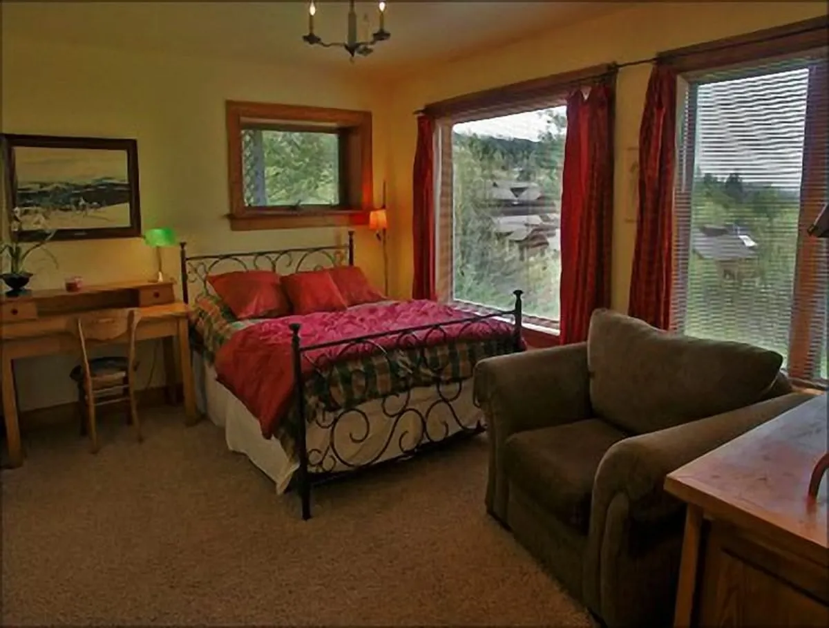 A spacious bedroom is shown with a large bed featuring decorative bedding, flanked by a comfortable armchair and a desk with a chair near large windows that provide scenic views. The room is bathed in natural light, creating a calm and restful atmosphere.