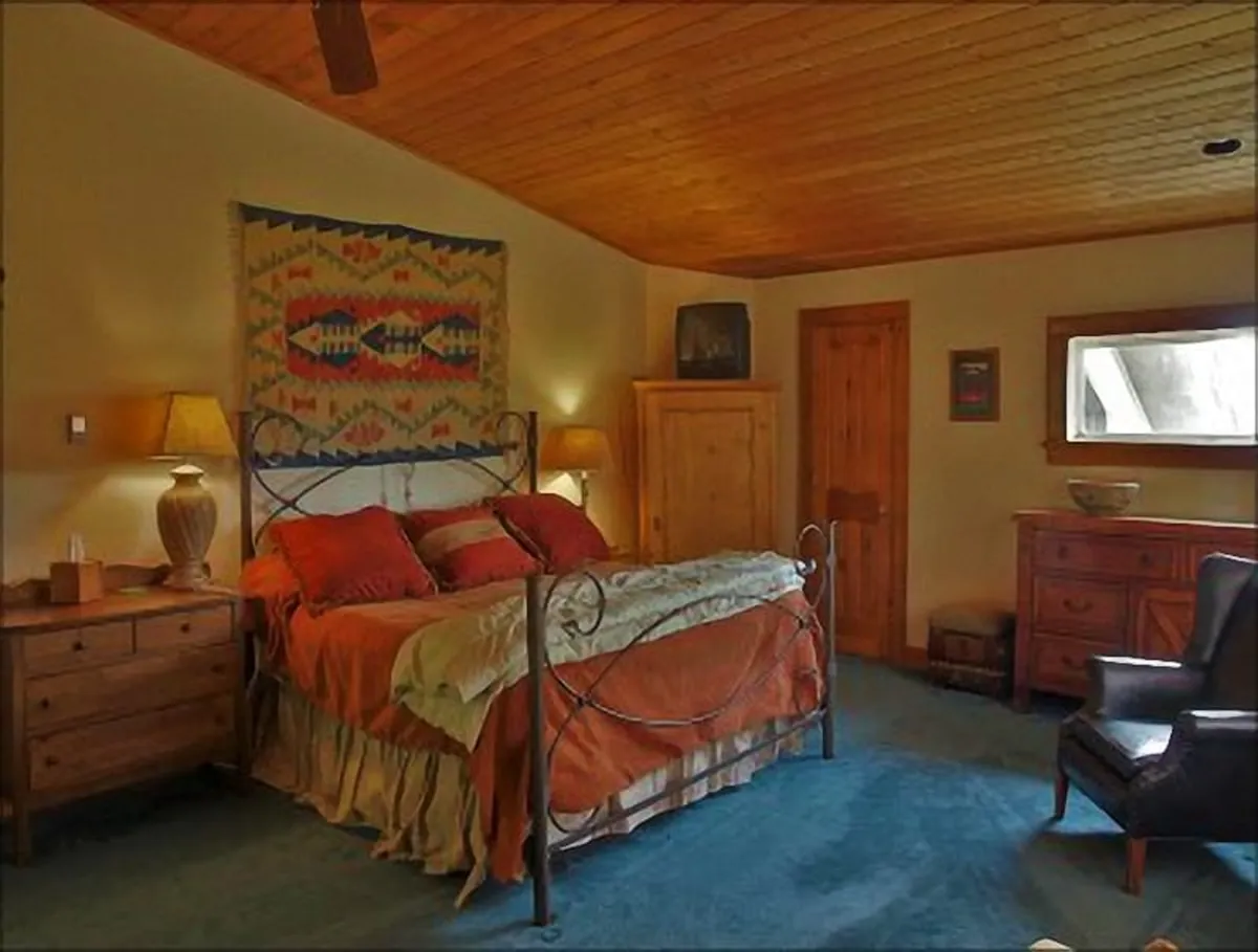 This image features a cozy bedroom with a neatly made bed, decorative pillows, a vibrant wall hanging, and warm wooden furniture. Natural light filters into the space, creating a welcoming and restful atmosphere ideal for unwinding.