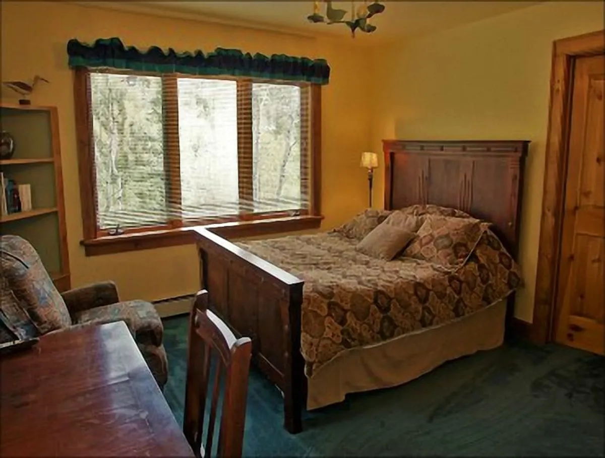 A sunlit bedroom with large windows that frame a view of nature, featuring a wooden bed and a comfy armchair beside a desk. The space feels peaceful and connected to the outdoors, offering a spot to unwind, read, or plan the next day's activities while enjoying the scenery.