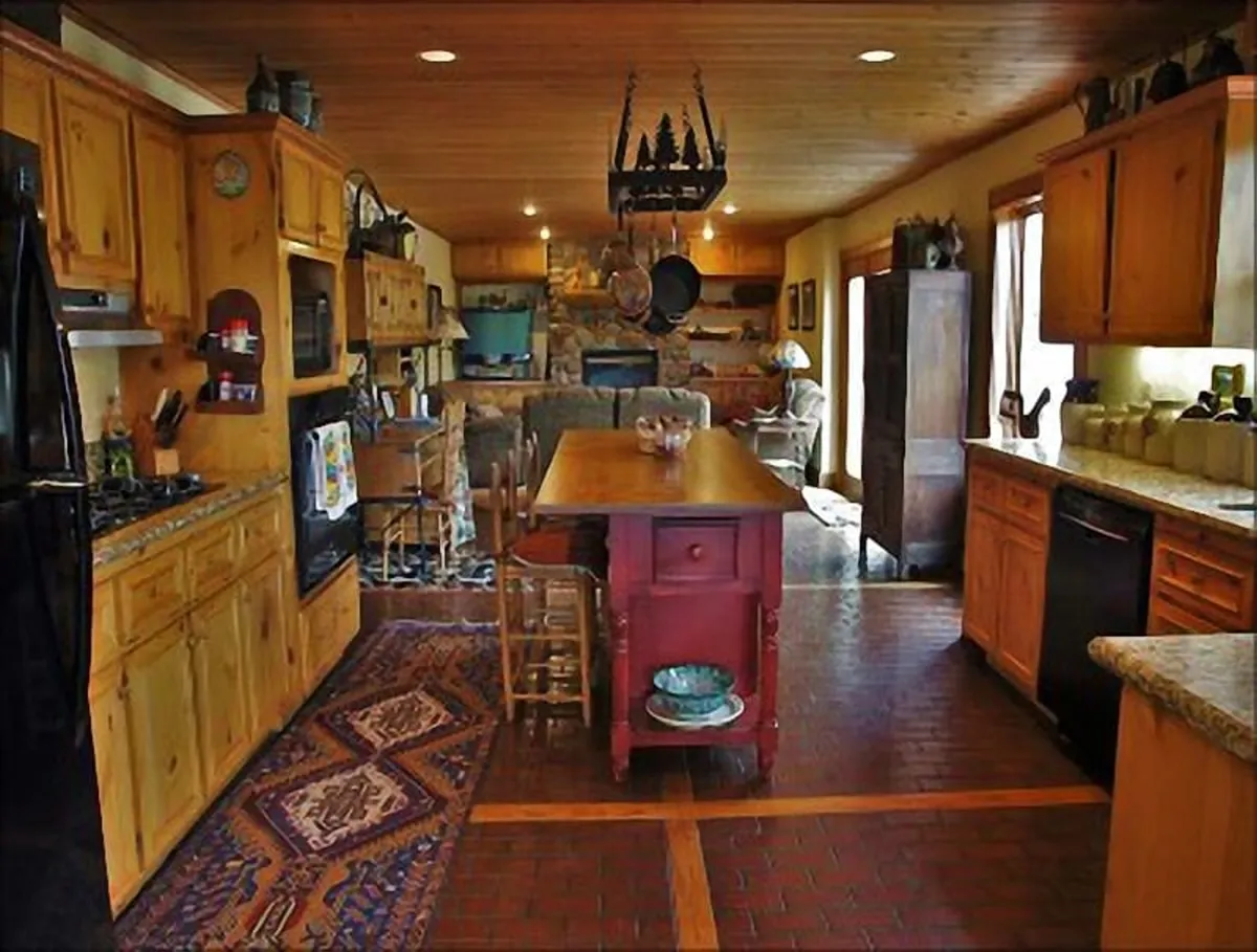 Here, the focus is on a large central kitchen island, wood cabinetry, brick flooring, and shelves adorned with decorative items. Kitchen tools hang from a rack, signaling a space designed for group meal prep and baking. The setting exudes a cozy, homey feeling, inviting everyone to participate.